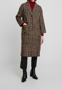 Cappotto lungo a quadri con una miscela di tonalità marroni, nere e rosse, caratterizzato da un colletto, due tasche anteriori e una chiusura con bottoni.