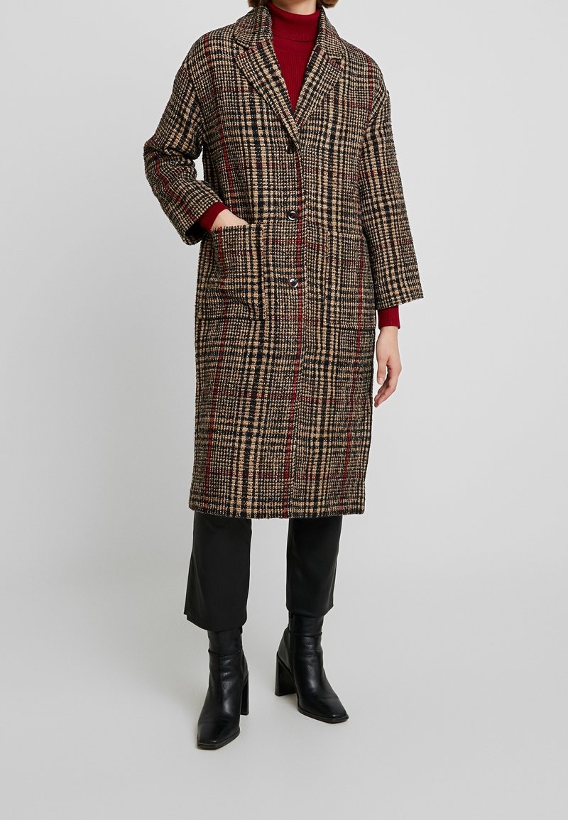 Cappotto lungo a quadri con una miscela di tonalità marroni, nere e rosse, caratterizzato da un colletto, due tasche anteriori e una chiusura con bottoni.