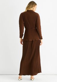 Brown pleated maxi dress with long sleeves, waistband detailing, textured fabric, and a flowy silhouette; worn with light-colored heels.