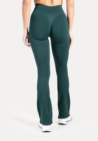 Vrouw die een donker groene legging met hoge taille en witte sneakers draagt, van achteren gezien tegen een effen witte achtergrond.