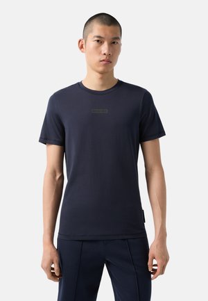 Marineblaues Baumwoll-T-Shirt mit Rundhalsausschnitt, kurzen Ärmeln und einem dezenten Logo in der Mitte. Glatte Textur und taillierter Schnitt, kombiniert mit dunklen Hosen.
