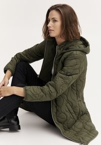 Olivza zaļa quilted jaka ar kapuci, rāvējslēdzi un melno akcentu detaļām. Modelis valkā melnus bikses un sēž pret vienkāršu fonu.