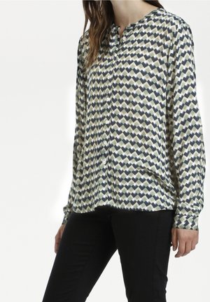 Femme portant une chemise boutonnée à manches longues avec un motif géométrique et un pantalon noir, debout les bras détendus le long du corps.