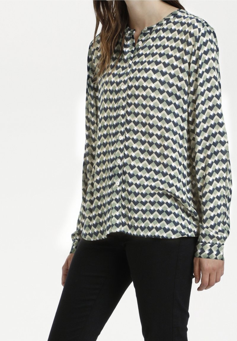 Femme portant une chemise boutonnée à manches longues avec un motif géométrique et un pantalon noir, debout les bras détendus le long du corps.