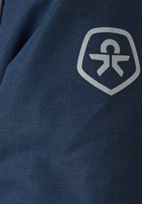 Granatowy materiał z teksturą, z srebrnym logo w kształcie tarczy z stylizowaną postacią w jej centrum.