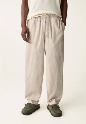Homme portant un pantalon beige à cordon, un haut sans manches en mesh blanc et des chaussures slip-on vert olive, debout les mains dans les poches.