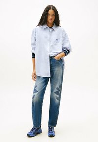 Mujer de pie, vestida con una camisa de rayas azul y blanca de gran tamaño, jeans bordados en azul con patrones florales y zapatillas deportivas azules.