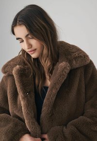 Brun faux fur frakke med en bred krave og blød tekstur. Har oversized design og subtile metal detaljer, der giver varme og komfort.
