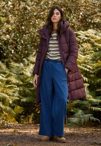 Bordeaux gekleed gewatteerde jas met capuchon, gestreept shirt, wijde blauwe denim broek en tan klompen, tegen een natuurlijke, groene achtergrond.