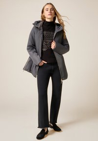 Grå puffjakke med hætte, sort mønstret sweater under, mørkeblå jeans og sorte teksturerede slip-on sko.