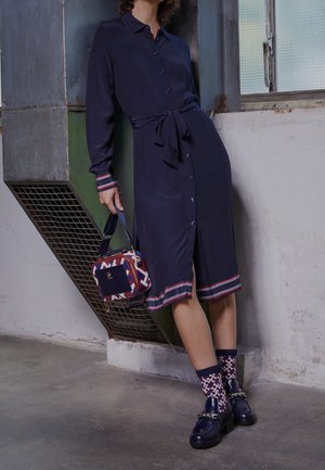 Robe chemise bleu marine avec col, taille ceinturée, poignets rayés. Associée à un sac à motifs et des chaussettes à motifs. Des mocassins épais complètent le look.
