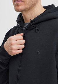 Čierna mikina na zips s kapucňou na sťahovanie, s hladkým povrchom a malým logom na hrudi. Zips je viditeľný.