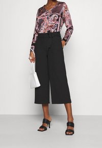 Langermige blouse met kleurrijk paisleypatroon, gecombineerd met hoge zwarte wijde broek en zwarte haksandalen, terwijl er een witte tas wordt vastgehouden.