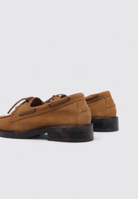 Braune Wildlederschuhe mit runden Zehen, Schnürdesign und dunklen Holzabsätzen. Glatte Textur mit sichtbaren Nähten und Ösen-Details.