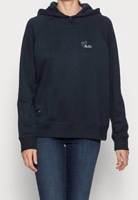 Donna che indossa un felpa blu navy con un piccolo cuore e un logo in corsivo sul petto, abbinata a jeans blu scuro, in piedi davanti a uno sfondo bianco.