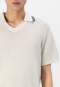 Šviesiai smėlinis v-neck megztas polo marškinėliai su tekstūruotu audiniu, jūrų laivyno juostelės akcentais ant apykaklės ir trumpomis rankovėmis.