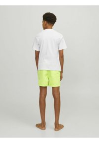 Camiseta blanca de manga corta y pantalones de baño verdes neón, con bolsillos traseros y un diseño decorativo de costura, vistos desde la parte trasera.