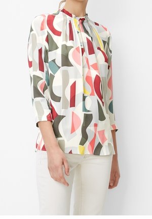 Vrouw draagt een blouse met lange mouwen met abstracte meerkleurige vormen en een witte broek, staand tegen een effen lichte achtergrond.