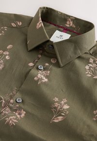 Oliivinvihreä paita, jossa kukkakuosi hillityissä vaaleanpunaisissa sävyissä, nappikauluksella ja harmailla napeilla, valmistettu sileästä kankaasta.