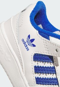 White sneaker with blue accents, featuring a textured tongue, zigzag patterned detailing, and an adidas logo in blue on the side.