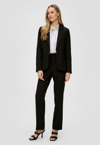 Zwarte op maat gemaakte pak met een getailleerd blazer en rechte broek. In combinatie met een witte gestreepte blouse en zwarte hoge hakken met bandjes.
