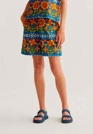 Mujer con pantalones cortos y top azules con un llamativo estampado floral rojo y naranja, combinados con sandalias azul marino, posando frente a un fondo liso.