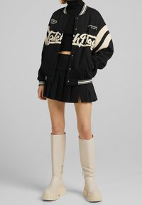 Schwarze Collegejacke mit cremefarbenen Akzenten, figurbetontem schwarzen Pullover und plissiertem schwarzen Rock, kombiniert mit knielangen beigen Plateau-Stiefeln.