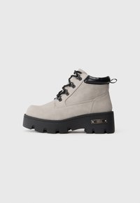 Botín de tobillo gris claro con cordones, suela negra gruesa, cuello acolchado negro y etiqueta de marca metálica en el lateral de la suela.