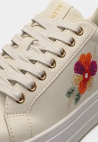 Baskets couleur crème avec de larges lacets blancs, œillets dorés et détail brodé de fleurs colorées sur le côté près de la semelle.