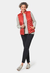 Rode gewatteerde vest met hoge kraag over een lichtgrijze trui. Zwarte broek en beige sneakers met witte veters. Slank, modern ontwerp.