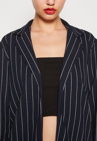 Námořnický blazer s bílými vertikálními pruhy, z měkké látky, se zářezovými klopami a otevřeným předním dílem. Nošený přes černý top bez ramínek, který zvýrazňuje linii krku.