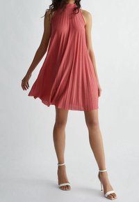 Donna che indossa un vestito a pieghe senza maniche di colore corallo e sandali con tacco alto bianchi, in piedi contro uno sfondo grigio chiaro uniforme.