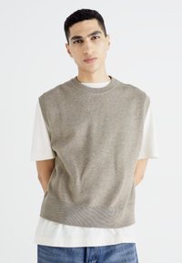 Pull sans manches marron clair avec ourlet et épaules côtelés, porté par-dessus un t-shirt blanc. Le modèle se tient debout, les mains sur les côtés, devant un fond uni.