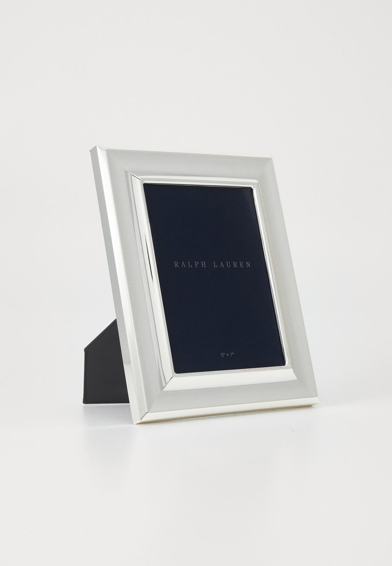 Cornice per foto rettangolare tono argento con un bordo riflettente, supporta uno sfondo blu scuro etichettato "RALPH LAUREN," di dimensioni 5x7 pollici.