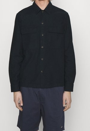 Mand iført en sort langærmet skjorte med knapper og to brystlommer samt mørkeblå knælange shorts.