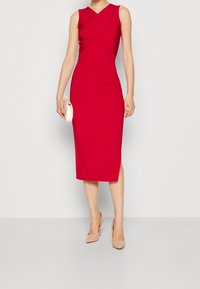 Červené bezrukávové šaty s přiléhavým střihem, zdobené křížovým designem na těle a sukní ke kolenům, dopln�ěné o nahé vysoké podpatky.