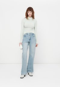 Haut à manches longues en ribbed vert clair associé à un jean évasé bleu clair et des chaussures de couleur crème. Le mannequin tient un petit sac blanc.