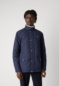 Jūras zilas quiltētas jakas ar augstu apkakli, spiedpogu un divām priekšējām kabatām. Iespējas šuvju raksts un neliels logotips uz krūtīm.