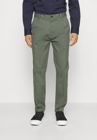 Pantaloni in cotone verde con vestibilità dritta, frontale piatto e due tasche laterali, abbinati a sneakers nere e una camicia blu scuro.