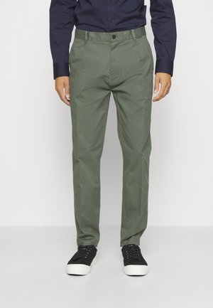 Pantaloni in cotone verde con vestibilità dritta, frontale piatto e due tasche laterali, abbinati a sneakers nere e una camicia blu scuro.