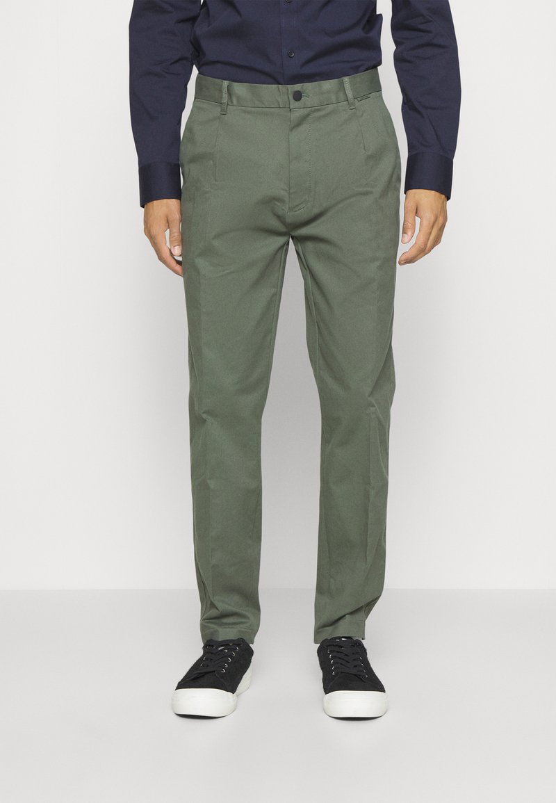 Pantaloni in cotone verde con vestibilità dritta, frontale piatto e due tasche laterali, abbinati a sneakers nere e una camicia blu scuro.