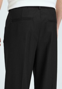 Pantalon noir avec une texture lisse, doté de deux poches arrière et d'une coupe ajustée. Lignes épurées et détails minimaux.