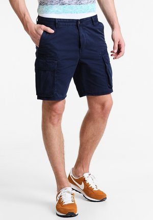 Mann trägt marineblaue Cargo-Shorts und orange-weiße Turnschuhe, steht mit einer Hand in der Tasche vor weißem Hintergrund.