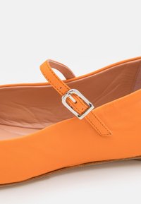 Chaussure plate en cuir orange avec une fine sangle ajustable et une boucle argentée, montrée en vue rapprochée de côté.