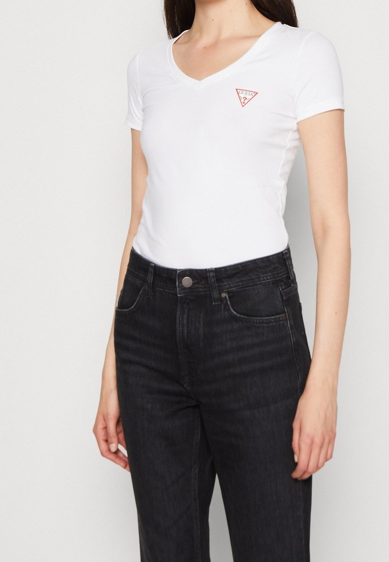 Femme portant un t-shirt blanc ajusté à col en V avec un petit logo rouge Guess et un jean noir taille haute, debout devant un fond uni.