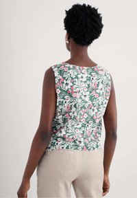 Cvetna brezrokavnik s zeleno podlago in rožnatimi, belimi ter mornarsko modrimi cvetličnimi vzorci; v kombinaciji s bež hlačami.