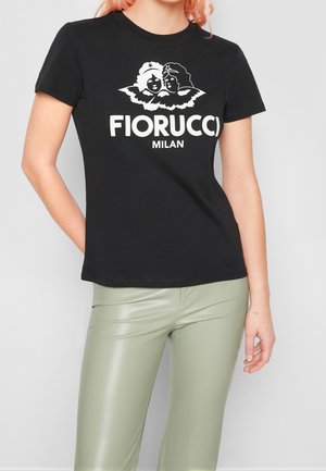 Zwart Fiorucci Milan T-shirt met witte afbeelding van twee engelen, gedragen met lichtgroene leren broek door een persoon met roze haar.