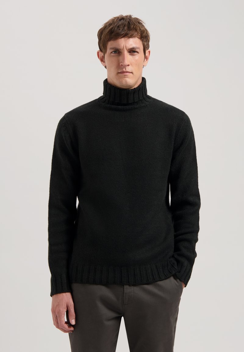 Schwarzer, gestrickter Rollkragenpullover mit gerippten Abschlüssen und langen Ärmeln, aus strukturiertem Stoff. Kombiniert mit dunklen Hosen.