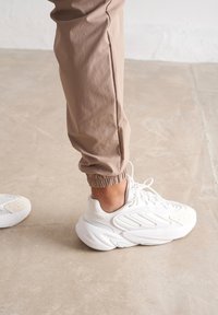 Baskets de sport blanches avec des revêtements texturés, des semelles courbées et des détails de laçage, associées à un pantalon de jogging beige clair.