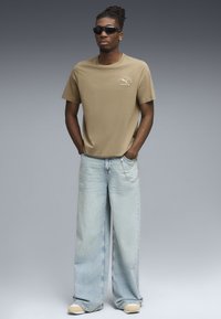 Korte mouwen katoenen T-shirt in tan met logo detail, gecombineerd met lichtblauwe wijde denim jeans en gele sneakers. Zwarte zonnebril gedragen.